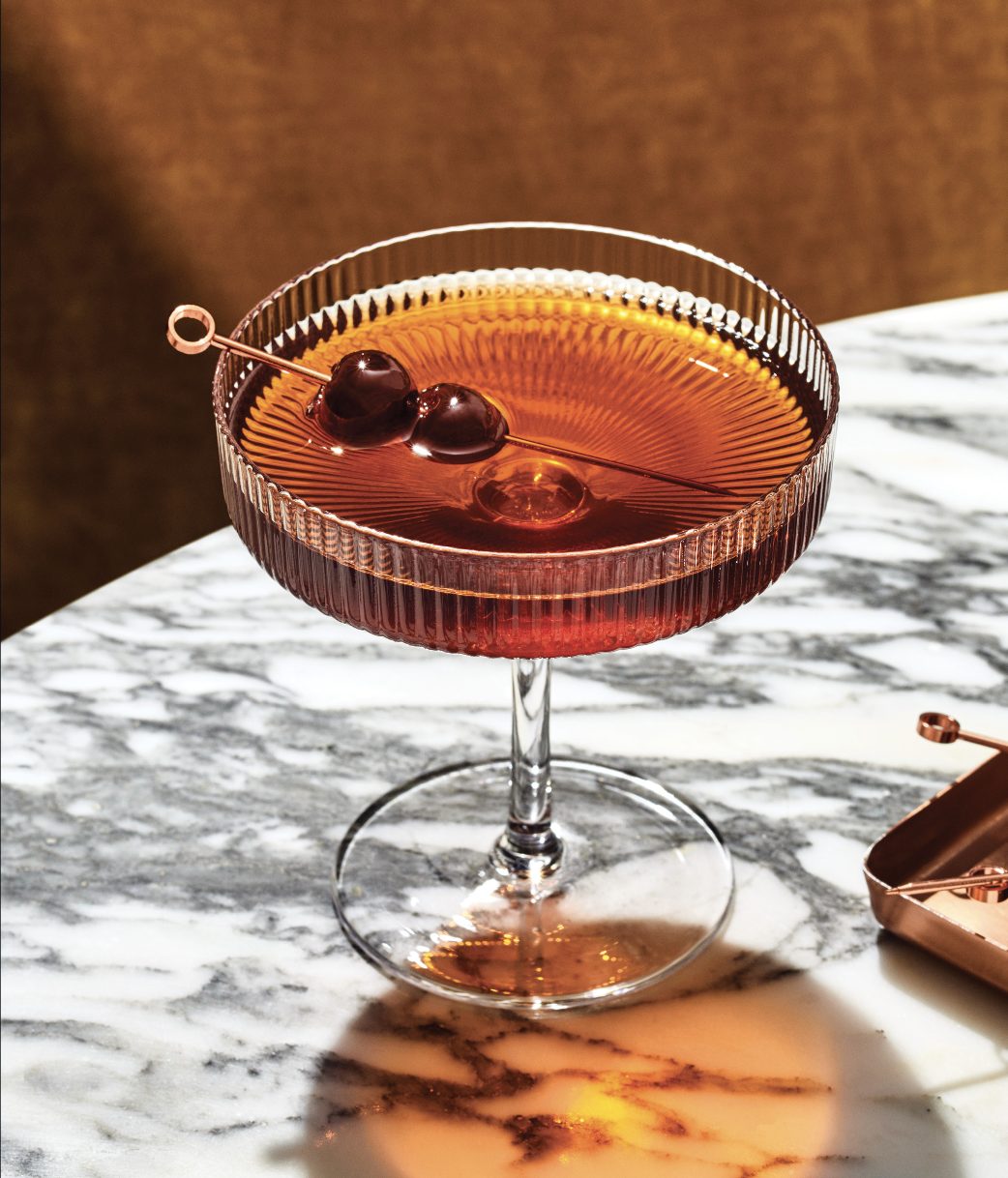 A Manhattan garnished with cherries in a ridged coupe on a marble counter.