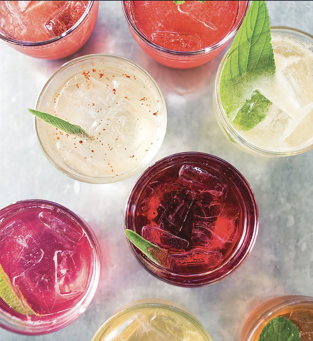 An overhead view of colorful ready-to-serve cocktails over ice with fresh garnishes.