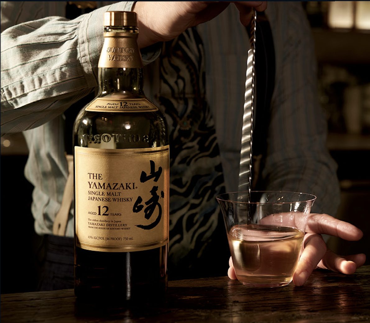 A bartender stirring a Yamazaki 12 Year Old whisky drink with a bar spoon beside the bottle.