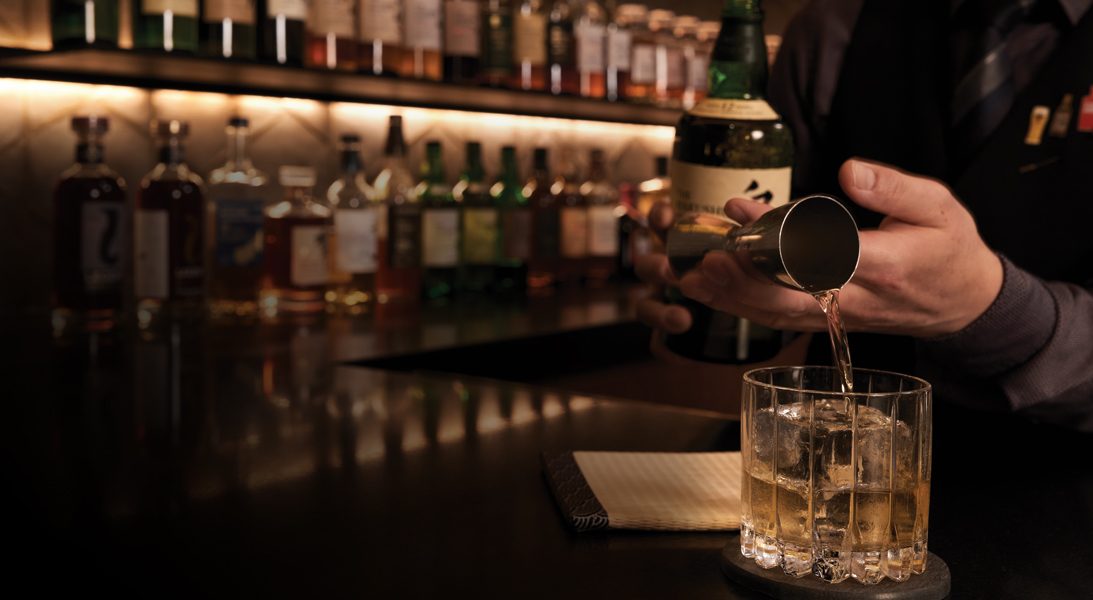 A bartender pouring a measured whisky over a large ice cube in a rocks glass.