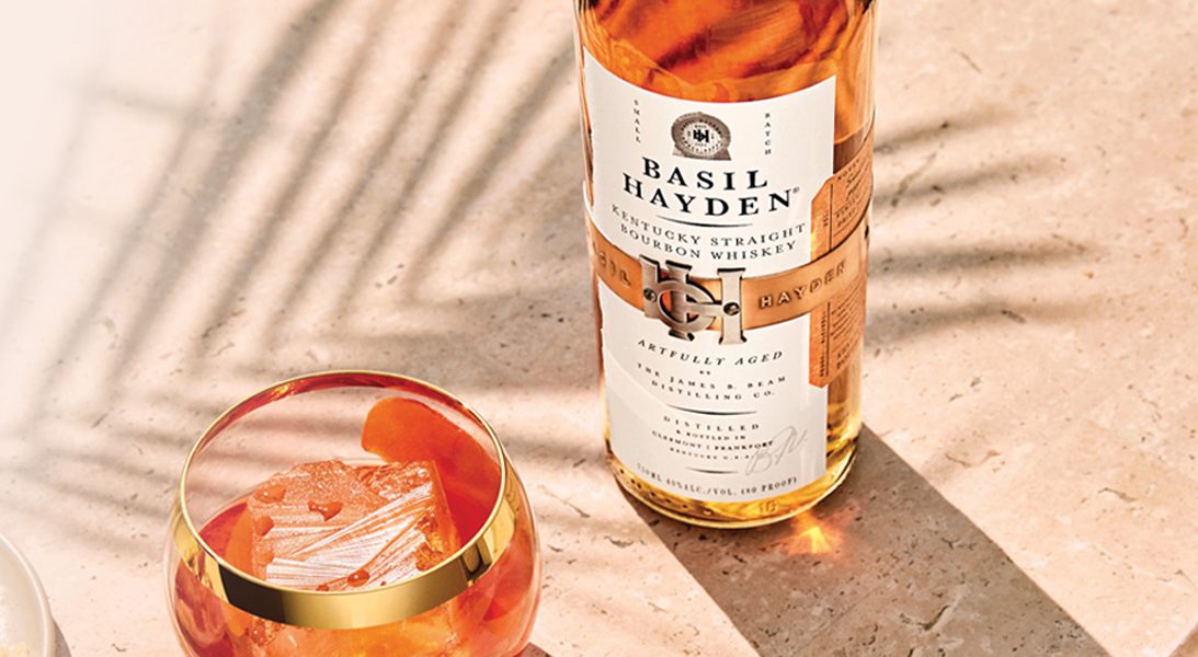 A bottle of Basil Hayden bourbon beside a cocktail over ice on a sunlit stone table.
